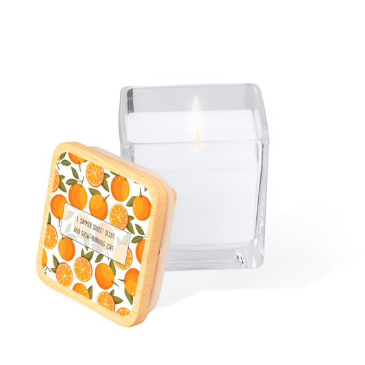 Vela aromática en tarro de cristal cuadrado con tapa de bambú. Diseño impreso: fondo color blanco con naranjas estilo acuarela. En el centro un rectángulo blanco con la frase: "A summer sunset scent and slow-burning love"