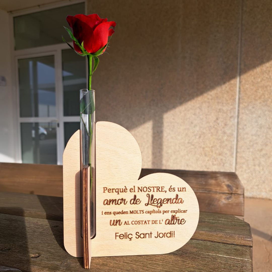 Porta rosas de madera formado por dos corazones cruzados y un tubo de cristal para poner la rosa con agua. Personalizado con una frase de amor grabada a láser en la madera especial para Sant Jordi. Frase: " Perquè el nostre, és un amor de llegenda i ens queden molts capítols per explicar un al costat de l'altre. Feliç Sant Jordi"
