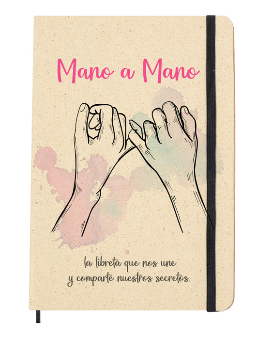 Portada del bloc de notas de hierba reciclada con goma elástica para mantenerla cerrada. Diseño de la portada: Dibujo de dos manos enlazadas por el dedo meñique, con manchas decorativas en rosa y morado.Acompaña la frase "Mano a mano, la libreta que nos une y comparte nuestros secretos.