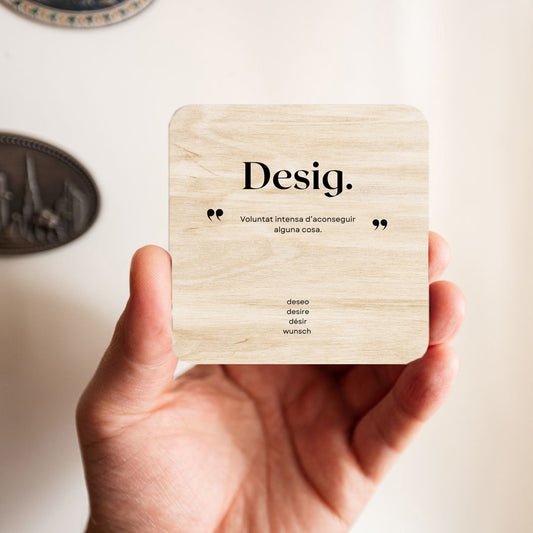 mano sujetando un imán de madera con la palabra DESIG y su definición y traducciones escrita en él. Forma parte de la colección Palabras Bonitas.