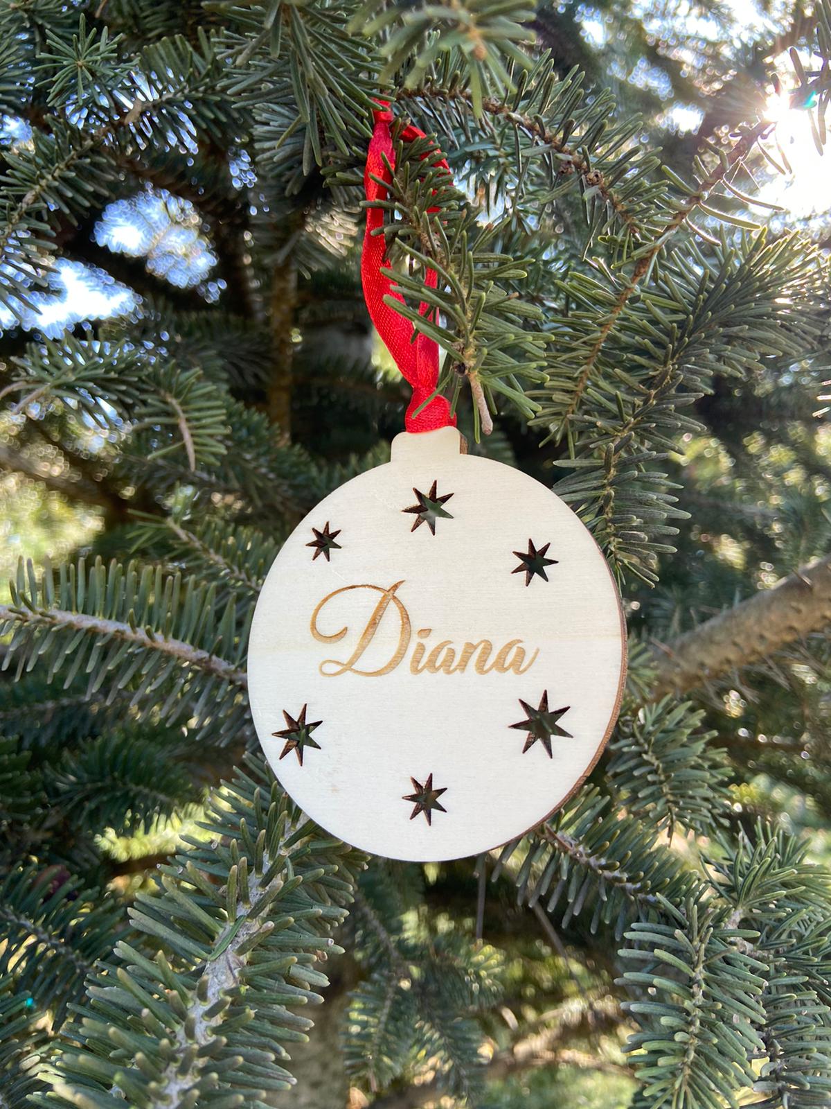 Bola para el árbol de Navidad con estrellas troqueladas y personalizada con el nombre grabado a láser. Incluye cinta roja para colgar.