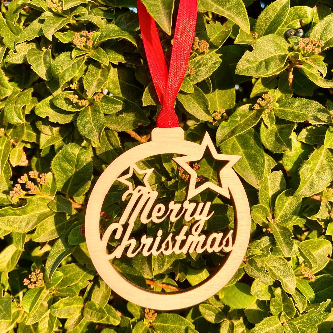 Bola de madera para el árbol de navidad con el mensaje Merry Christmas troquelado. Incluye cinta roja para colgar del árbol.