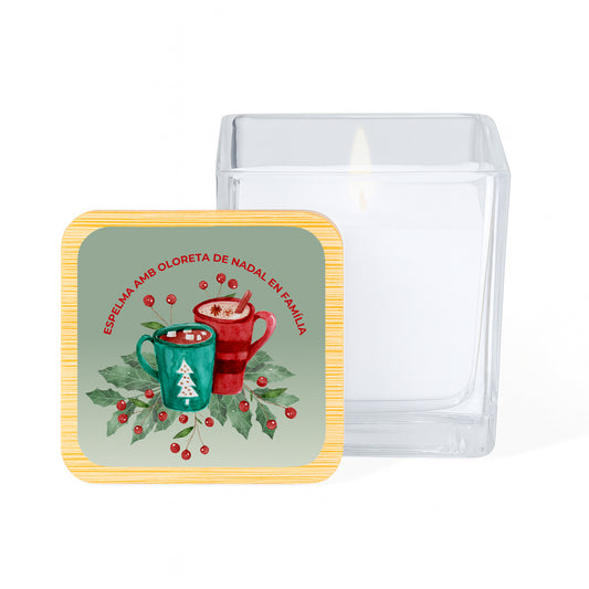 Vela con aroma de vainilla en tarro cuadrado de cristal y tapa de bambú impresa con un diseño de dos tazas de chocolate sobre fondo verde y la frase en rojo " Espelma amb oloreta de Nadal en Familia"