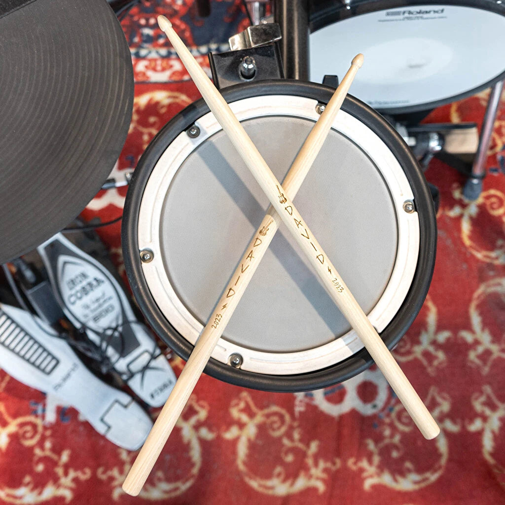 dos baquetas de madera personalizadas con un texto y fecha cruzadas sobre una batería profesional